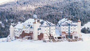 A large, multi-story building with a snow-covered roof and multiple chimneys, surrounded by a snowy landscape and dense forest. - Monte Baia Uludag (Bursa)