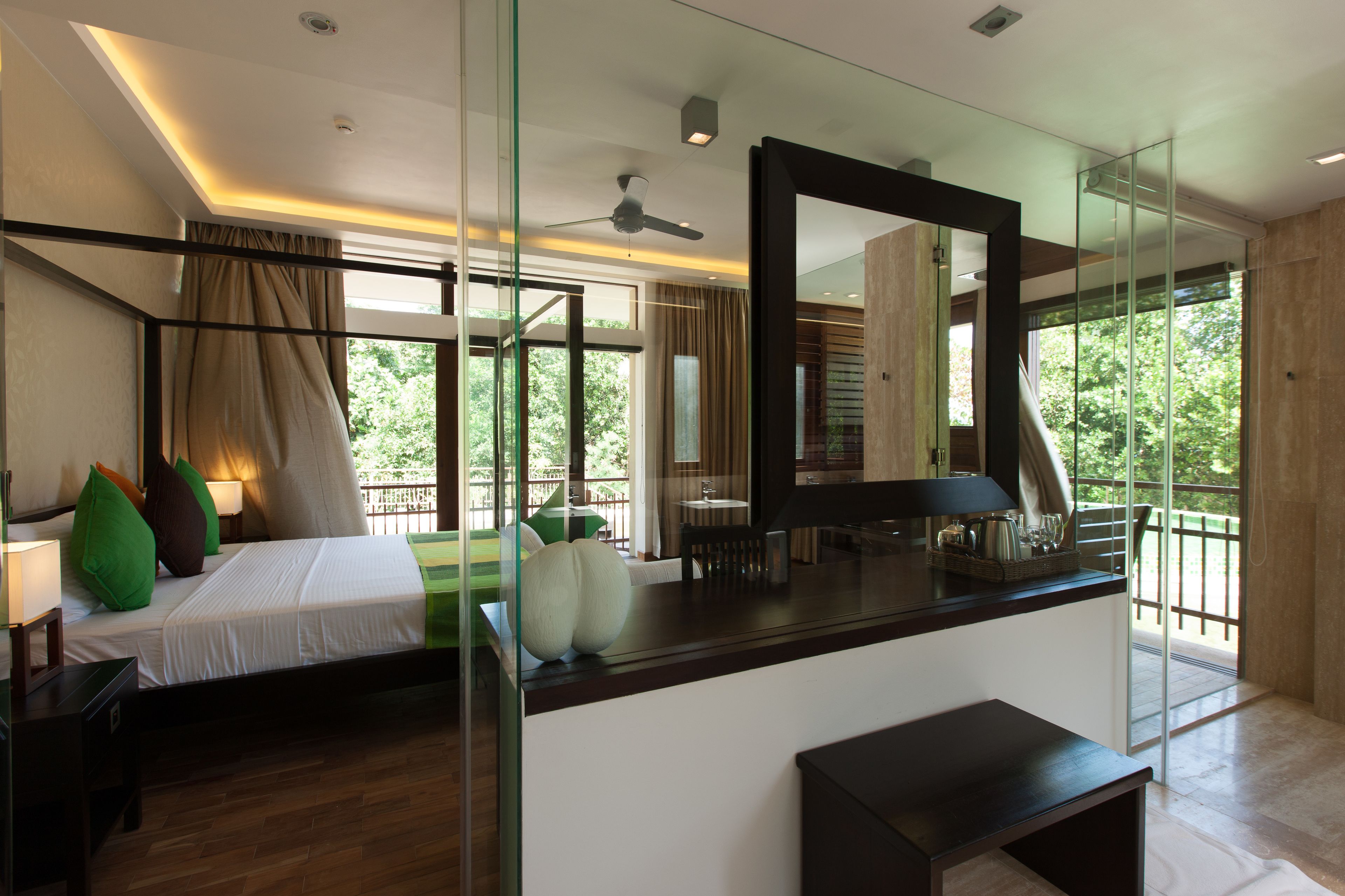 Una moderna camera d'albergo con un letto grande, una scrivania e un divisorio in vetro. Si nota una ventola a soffitto e vista sull'esterno.
