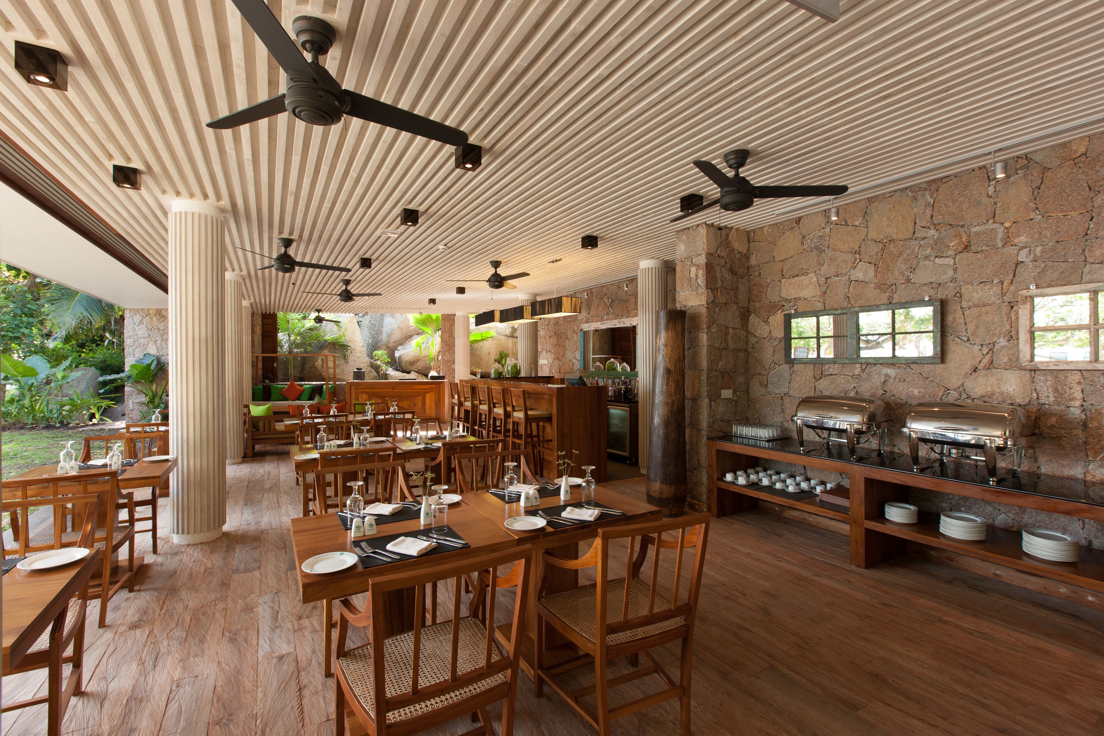 A spacious dining area with wooden tables and chairs, ceiling fans, and stone walls.