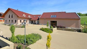 A building with a red roof and a beige facade, surrounded by a paved area with greenery and a clear sky.