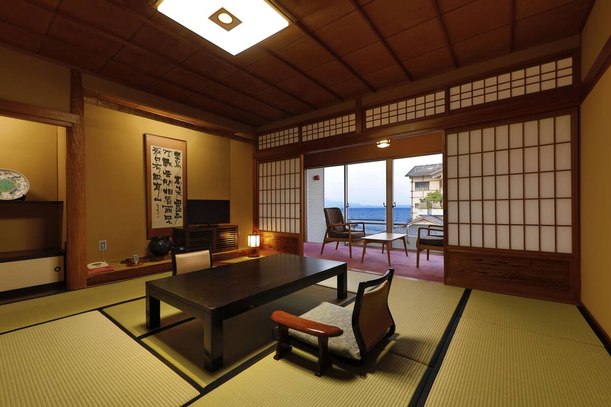 A traditional Japanese room with tatami flooring, a low table, and chairs. There is a view of the sea through a sliding door.
