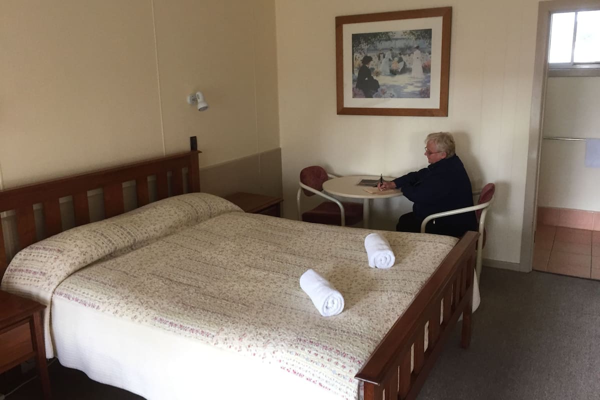 Hotellihuone, jossa on sänky, pieni pöytä, tuoli ja taulu seinällä.