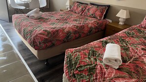 Et hotelværelse med to senge, begge med sengetæpper i et mønster af røde og grønne blade, en hvid stol, et lille bord og en lampe på et natbord.