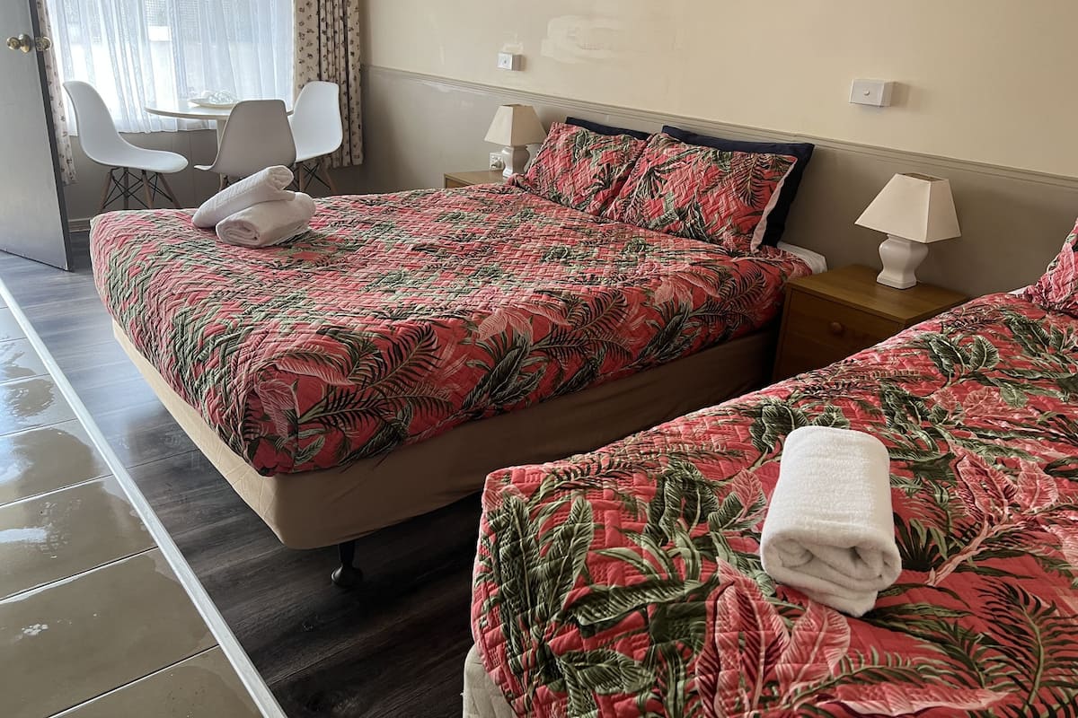 Hotellihuone, jossa on kaksi sänkyä. Jokaisessa sängyssä on punavihreäkuvioinen peitto, huoneessa on myös valkoinen tuoli, pieni pöytä ja yöpöydällä lamppu.