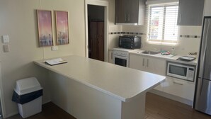 Et moderne køkken med en central ø, indbygget ovn, mikroovn og en brødrister. Der er et vindue med rullegardiner, en skraldespand og en dør til et andet rum.