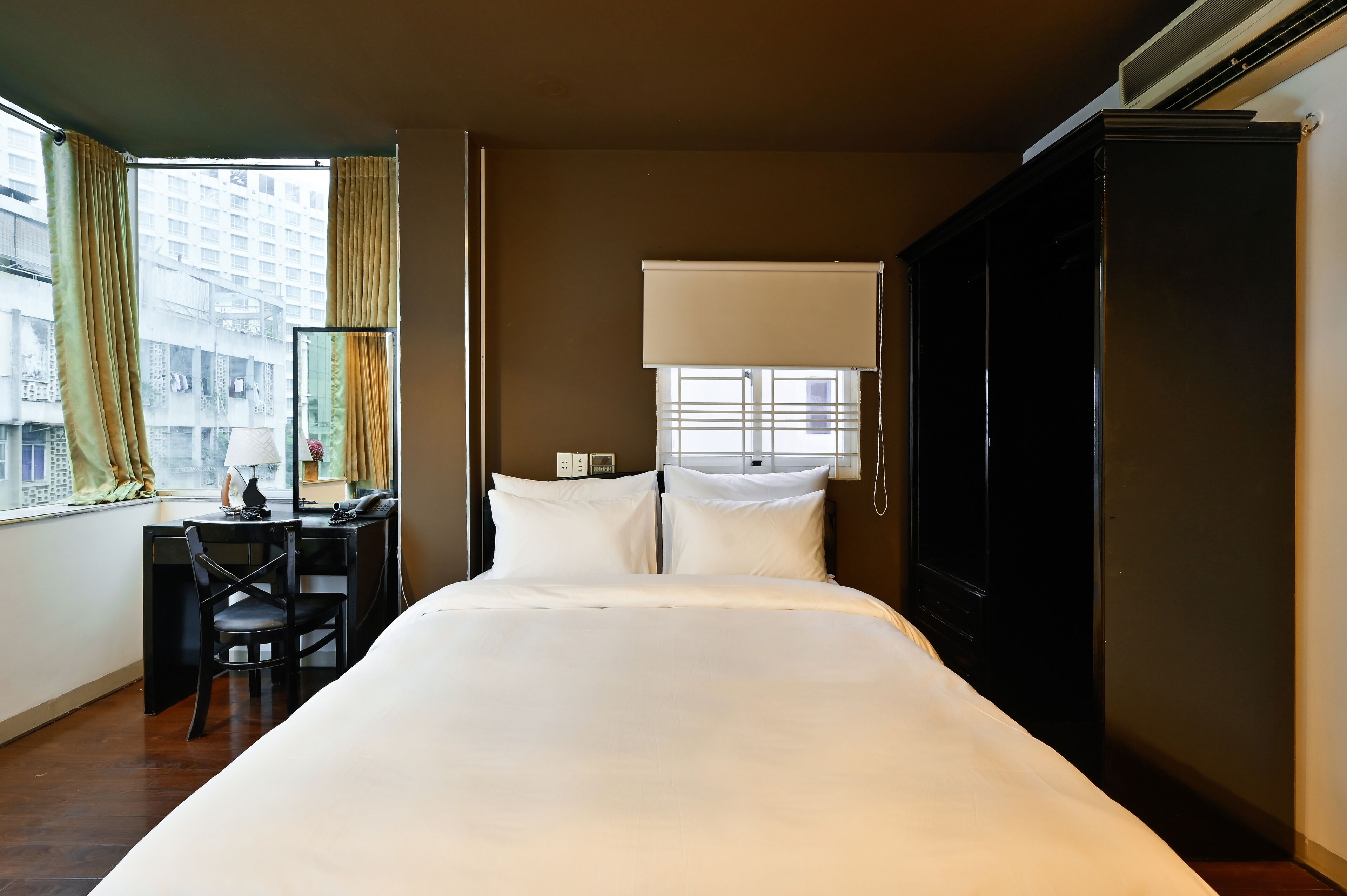 A hotel room with a bed, a desk with a chair, a window with curtains, and a wardrobe.