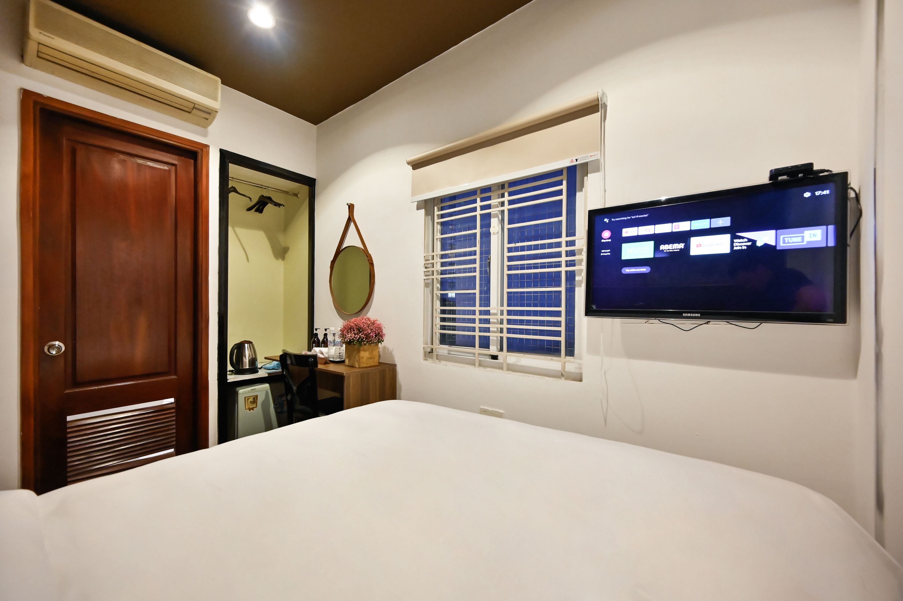 A hotel room with a bed, a television mounted on the wall, a window with blinds, a door, and a small table with a kettle and flowers.