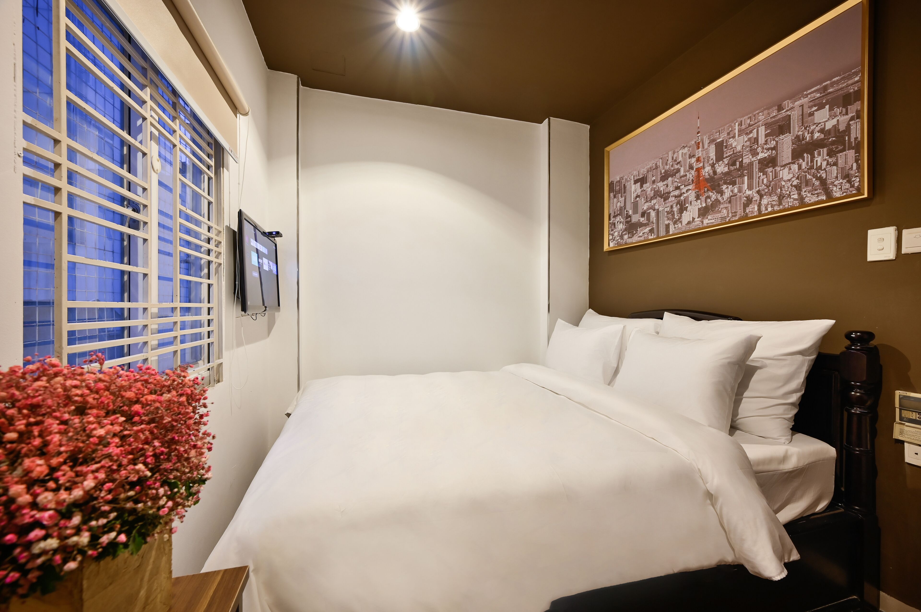 A hotel room with a bed, a window with blinds, a wall-mounted TV, a floral arrangement, and a cityscape painting.
