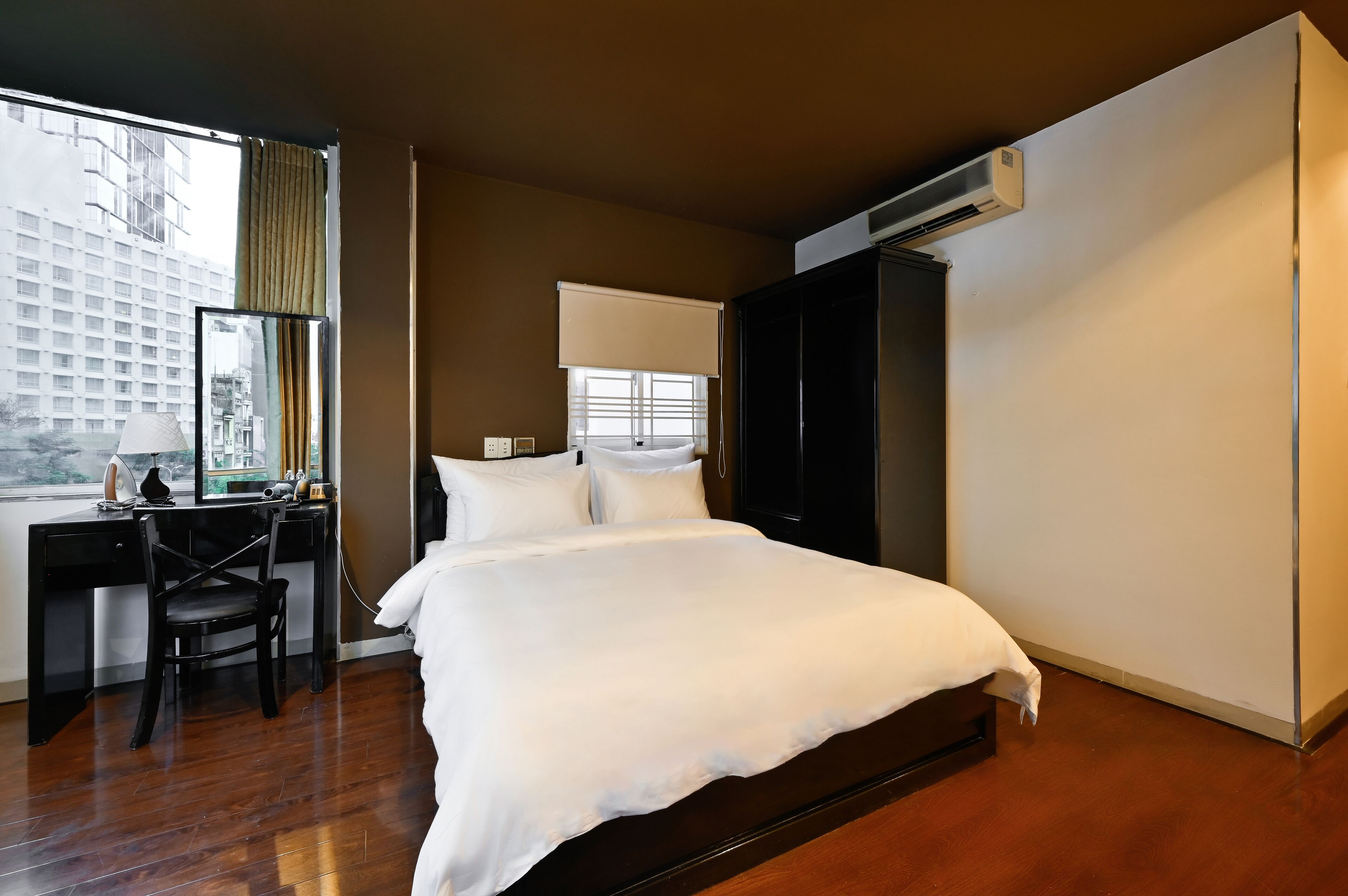 A hotel room with a large bed, a desk with a chair, a wardrobe, and an air conditioning unit.