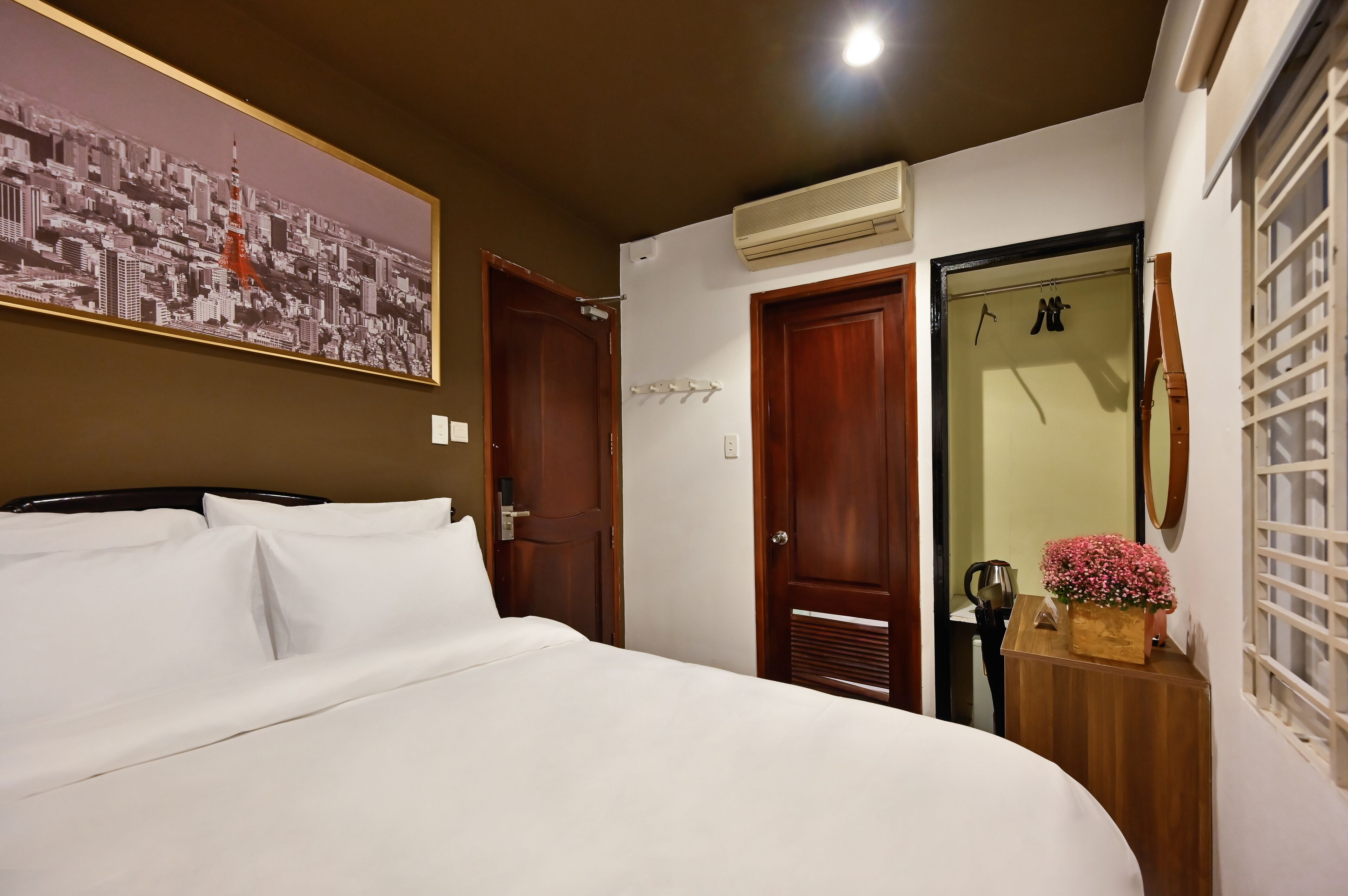 A hotel room with a bed, a wall-mounted air conditioner, a wooden door, and a small table with a flower vase.