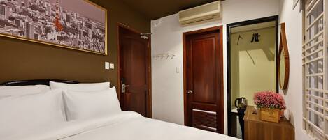 A hotel room with a bed, a wall-mounted air conditioner, a wooden door, and a small table with a flower vase.