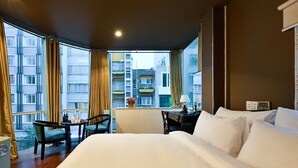 A hotel room with a large bed, a desk with a chair, a small table, and a view of a building through large windows. - The Noble Swan Hotel New World (Ho Chi Minh City)