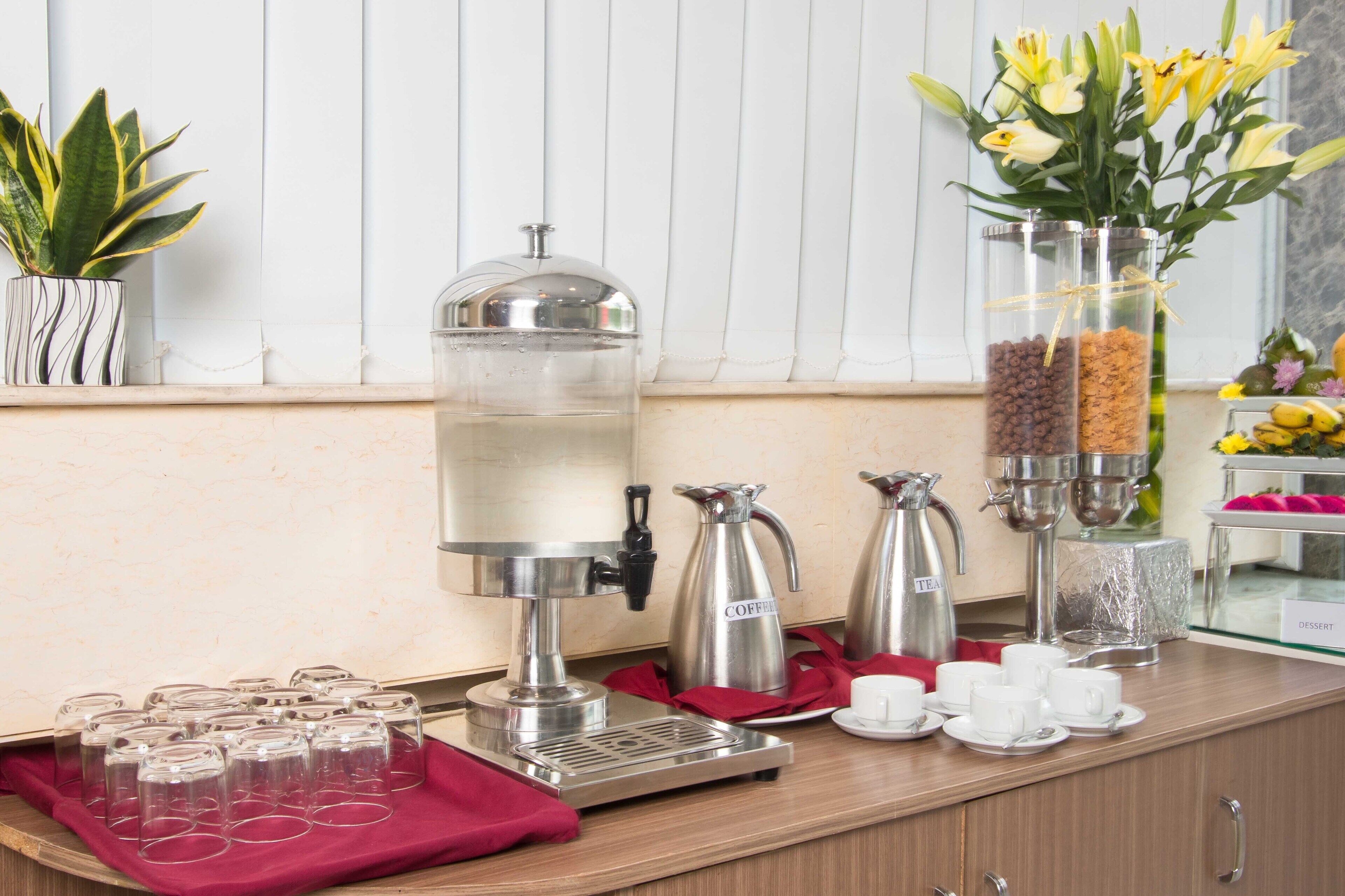 Una estación de café con dispensador de agua, cafeteras y dispensador de té. Hay tazas y platillos en una bandeja, y un jarrón con flores amarillas.