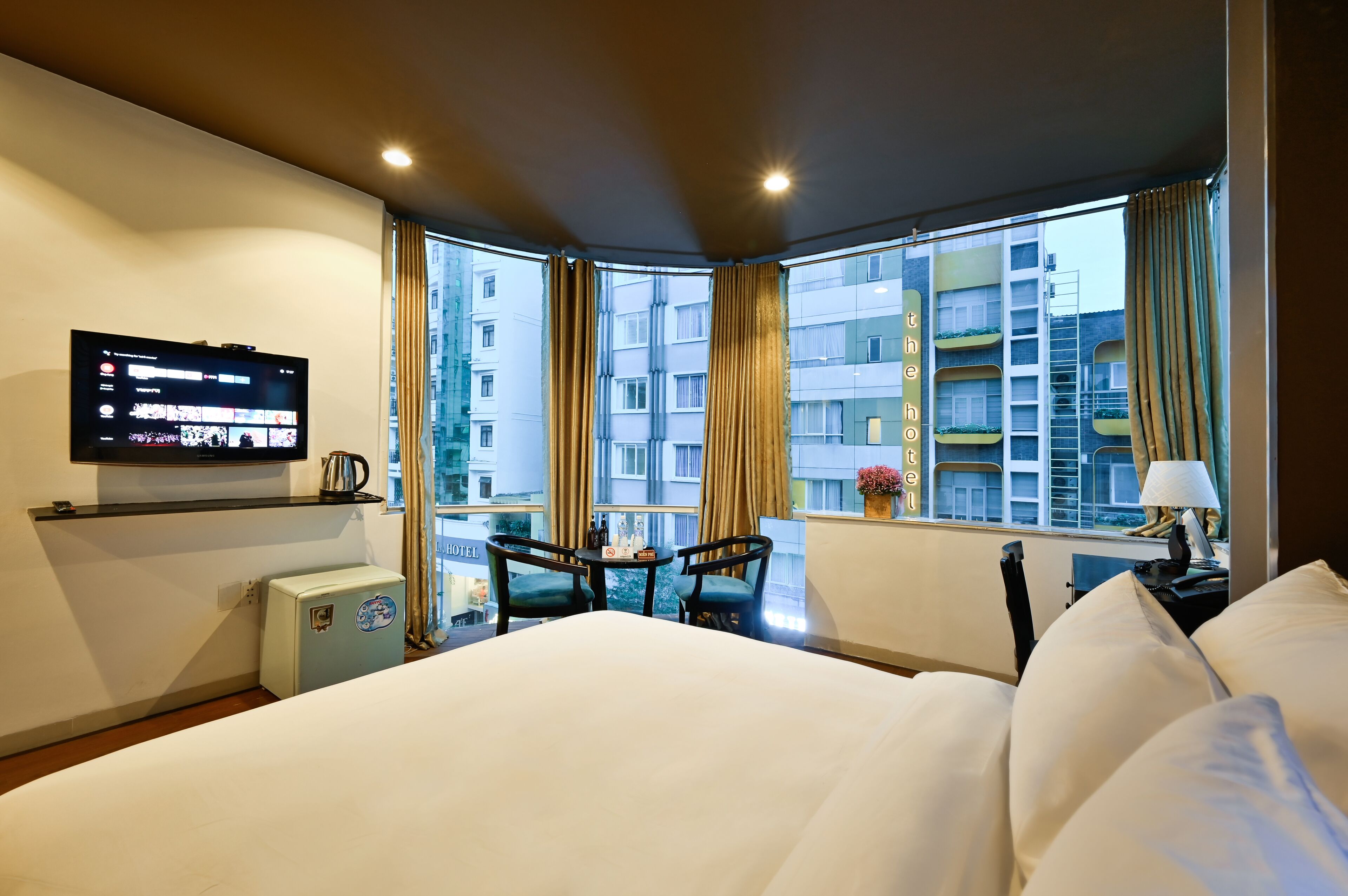 Una camera d'albergo con un letto, una TV fissata al muro, un tavolino con sedie e una finestra con tende.