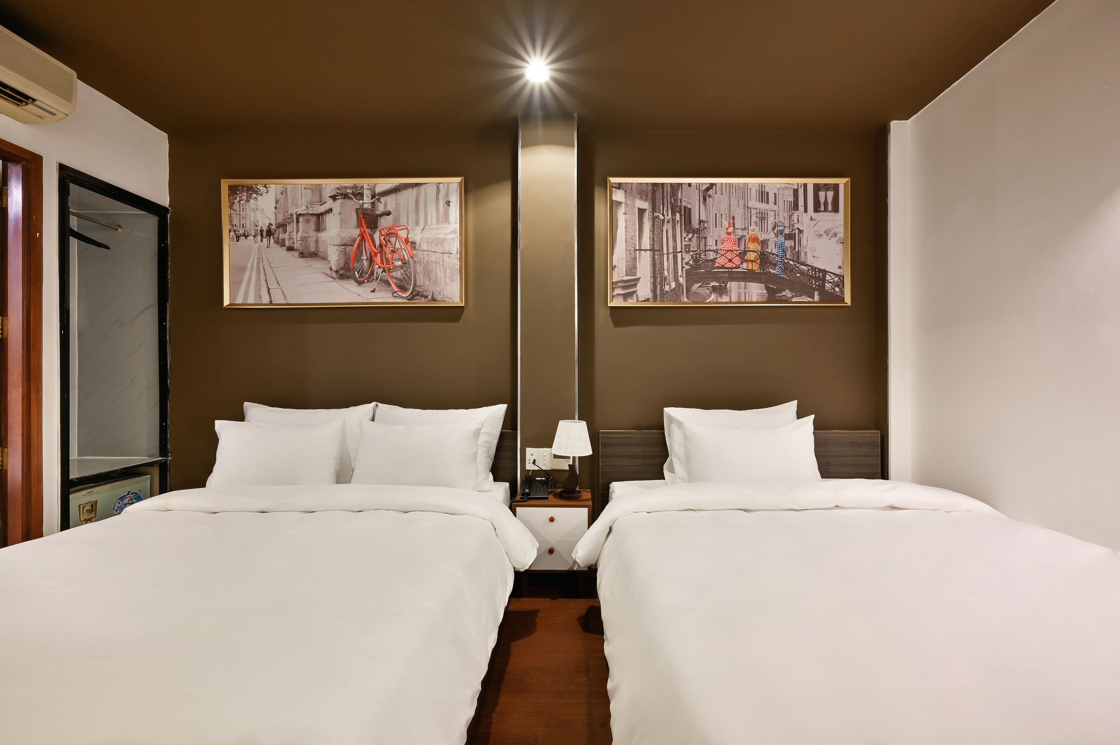 A hotel room with two beds, a nightstand, a lamp, and two framed pictures on the wall.