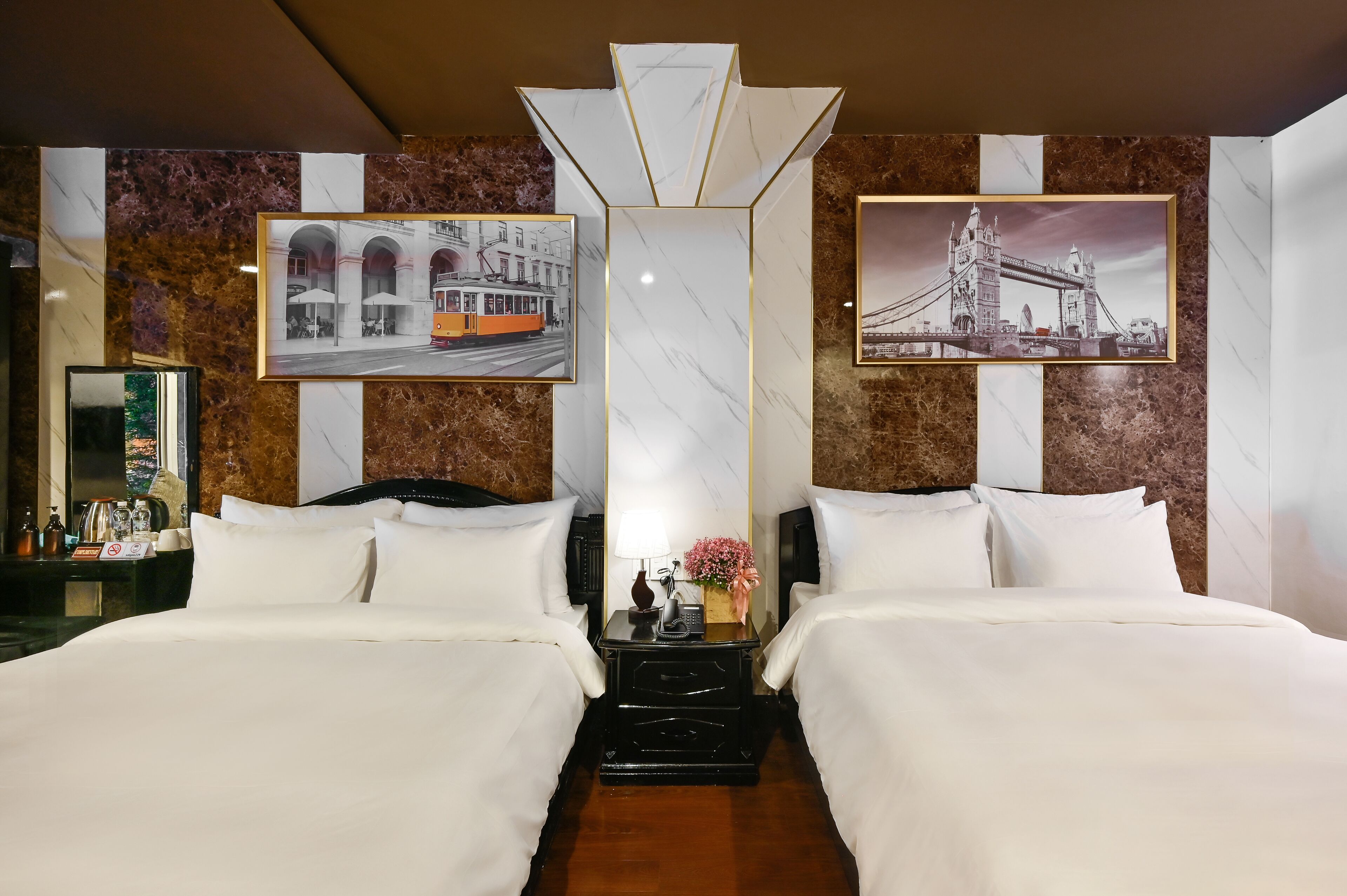 A hotel room with two beds, a nightstand with a lamp and flowers, and framed pictures of a tram and a bridge on the walls.