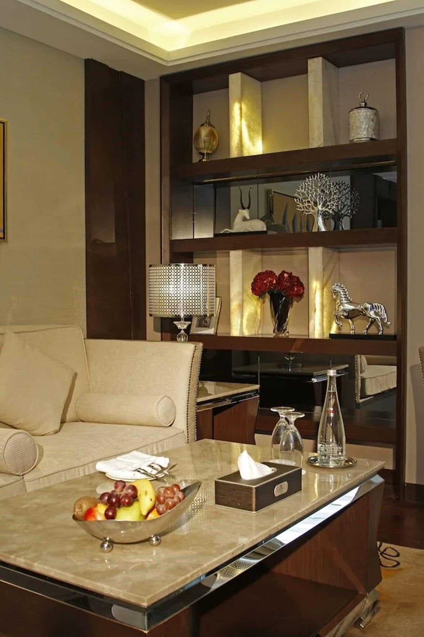 A modern living room with a sofa, a coffee table with fruits and a tissue box, and a decorative shelf with various items.