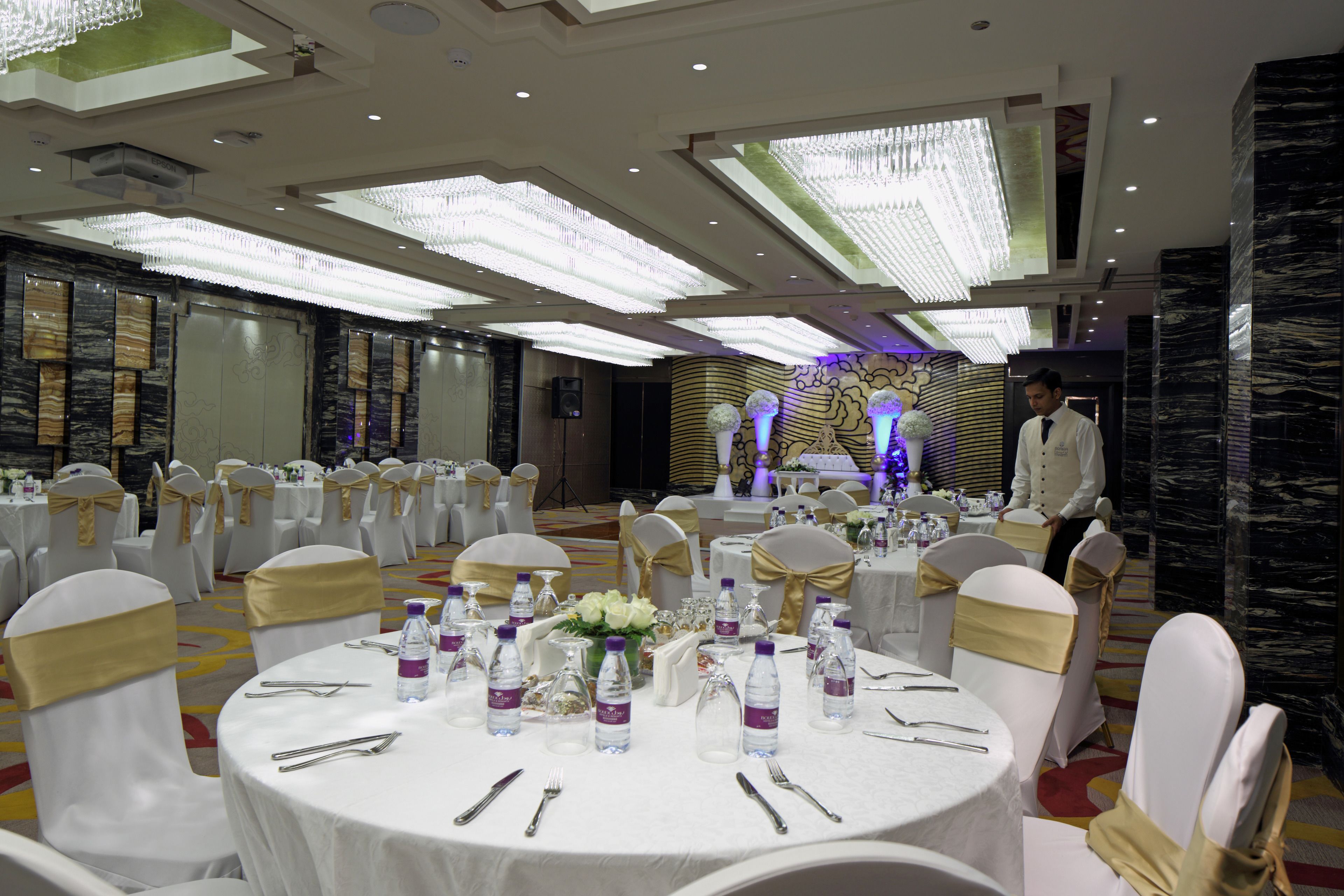 A banquet hall with round tables set for a formal event, featuring white tablecloths, gold sashes, and water bottles.