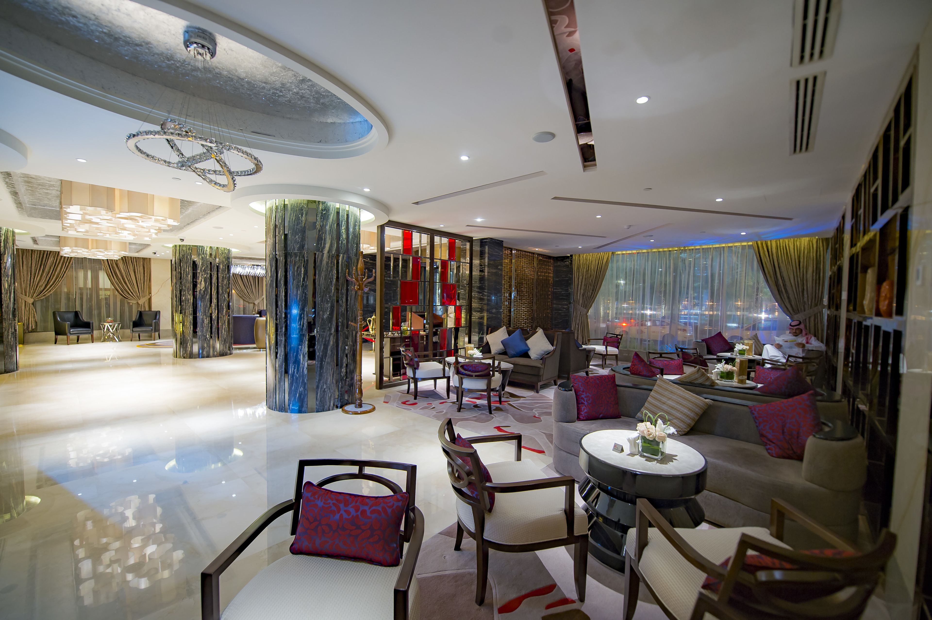 A modern hotel lobby with a central seating area, a chandelier, and a view of the cityscape through large windows.