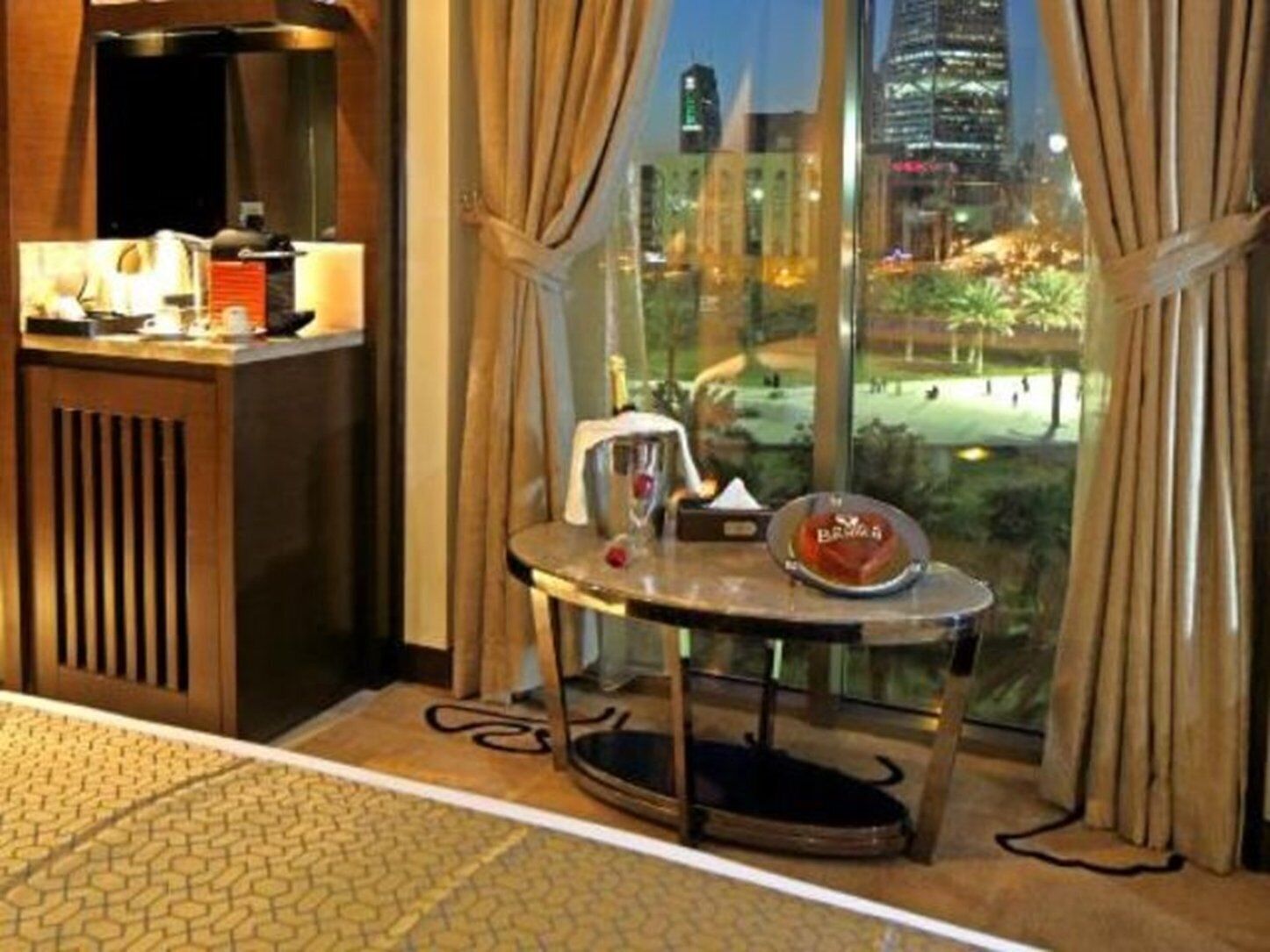 Habitación de hotel con vista a un paisaje urbano nocturno, una pequeña mesa con lámpara y una ventana con cortinas.