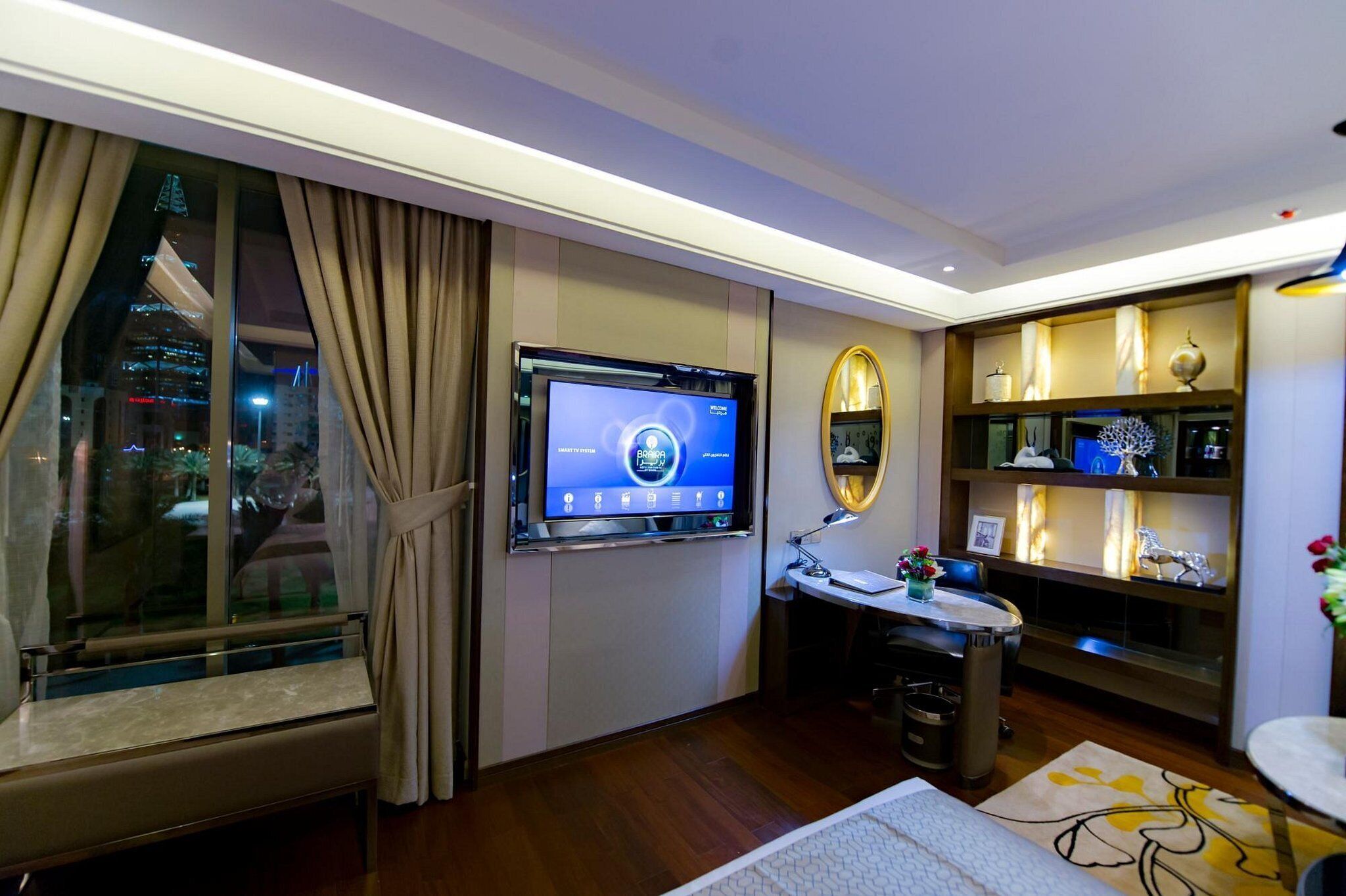 A hotel room with a large TV on the wall, a desk with a lamp, a small table with flowers, and a decorative shelf.