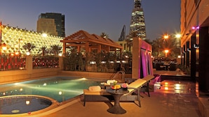 A rooftop pool area with lounge chairs, a small table with fruit, and a view of a city skyline at dusk. - Braira Hotel Olaya (Riyadh)