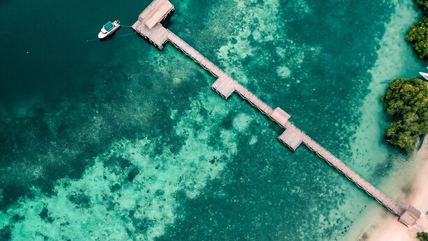 An aerial view of a beach with a wooden pier extending into turquoise waters, a swimming pool, and thatched-roof structures. - Sudamala Resort, Seraya, Flores (Labuan Bajo)