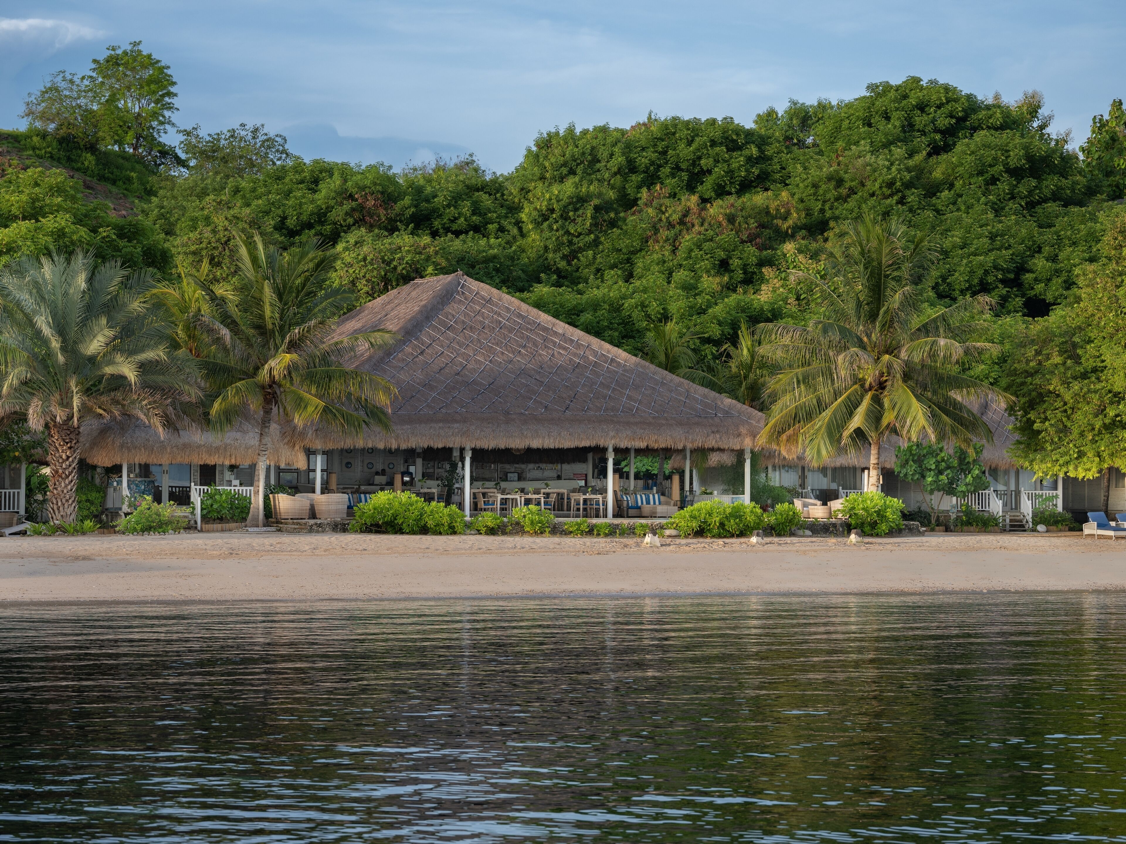 A beachside hut with a thatched roof, surrounded by lush greenery and palm trees.