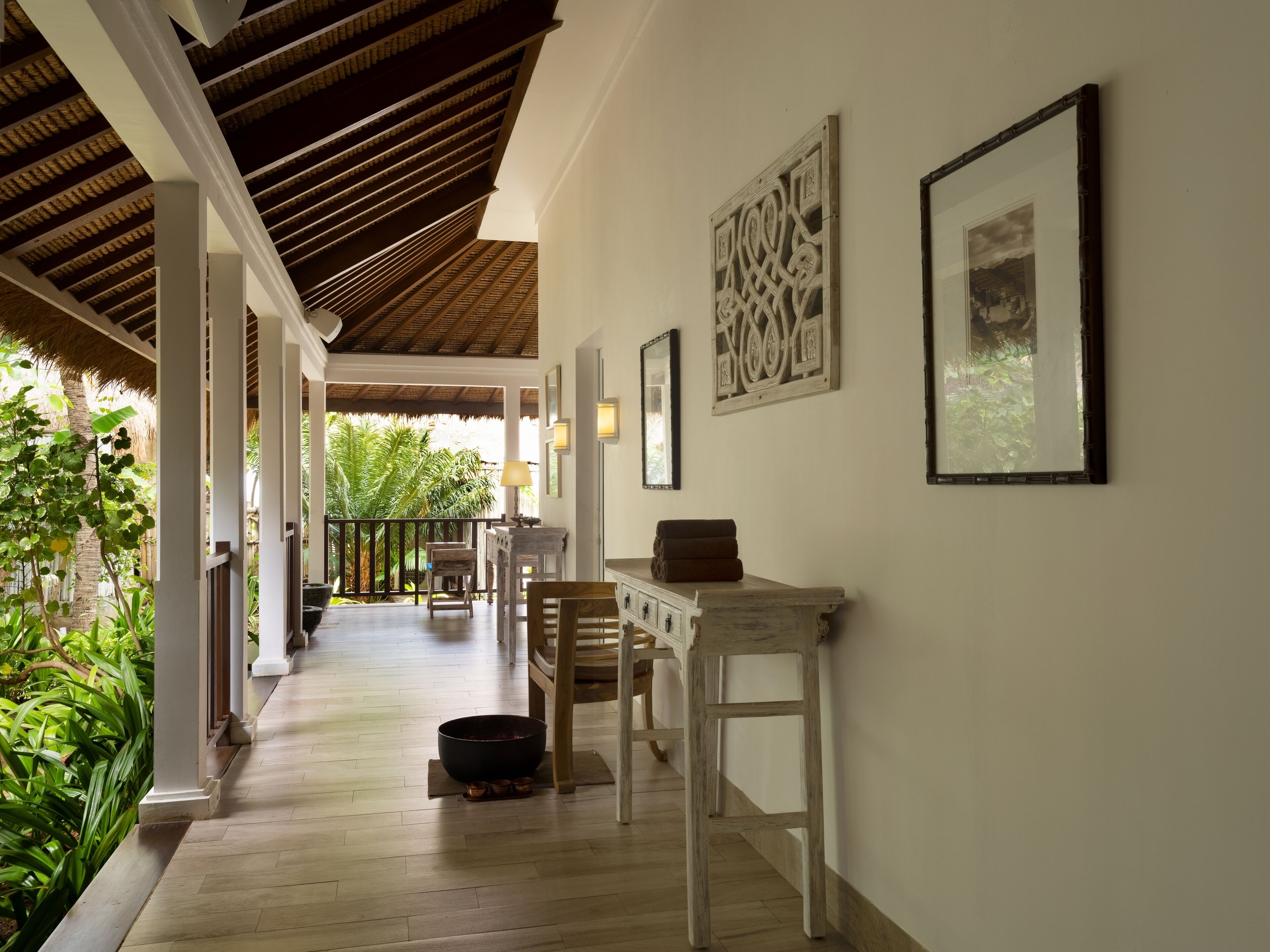 A corridor with a wooden floor, white walls, and a thatched roof. There are framed pictures on the wall and a table with a bowl and a towel on it.