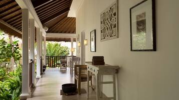 A corridor with a wooden floor, white walls, and a thatched roof. There are framed pictures on the wall and a table with a bowl and a towel on it.