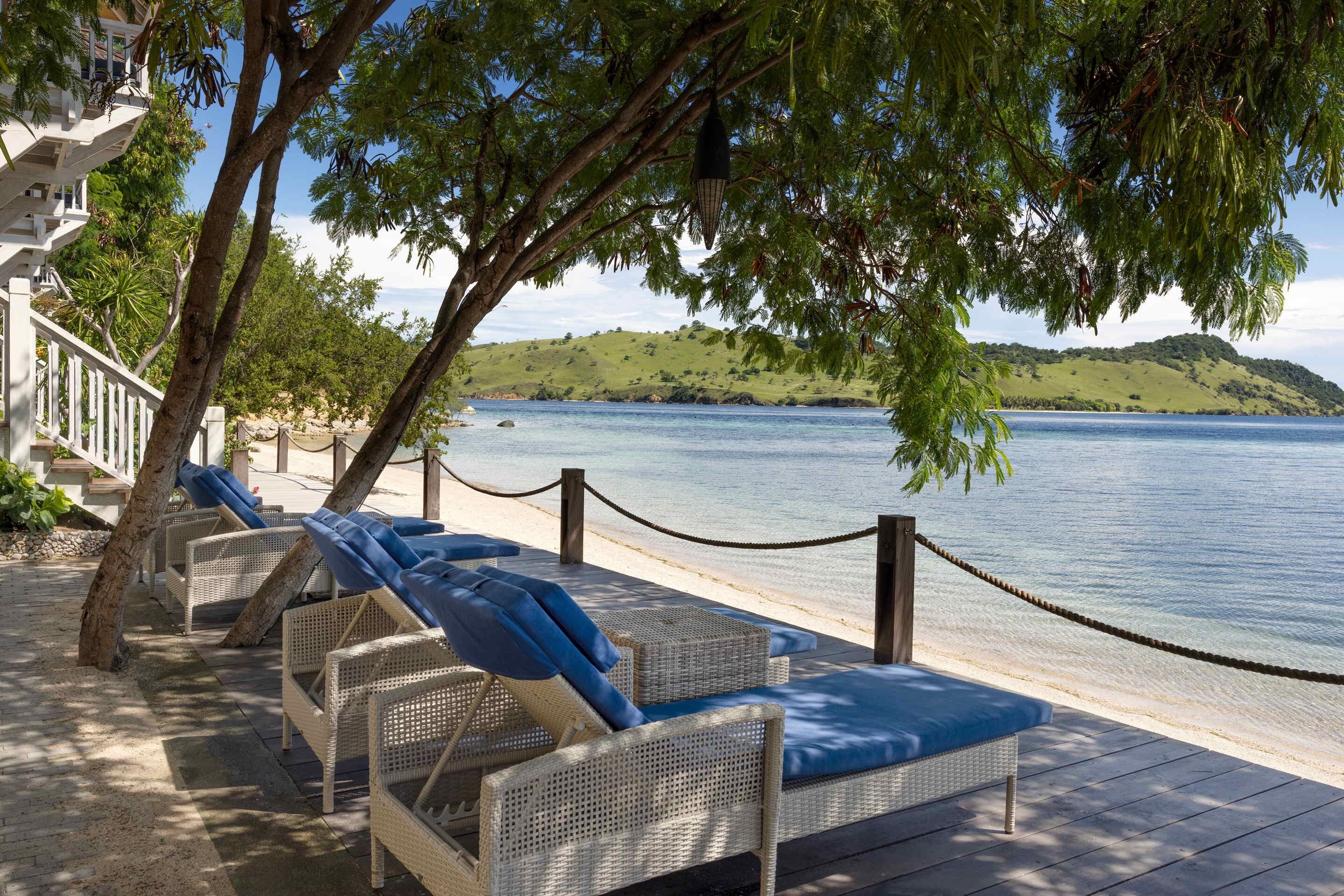 Eine Strandlounge mit Korbstühlen und blauen Kissen, mit Blick auf ein ruhiges Meer und einen grünen Hügel.