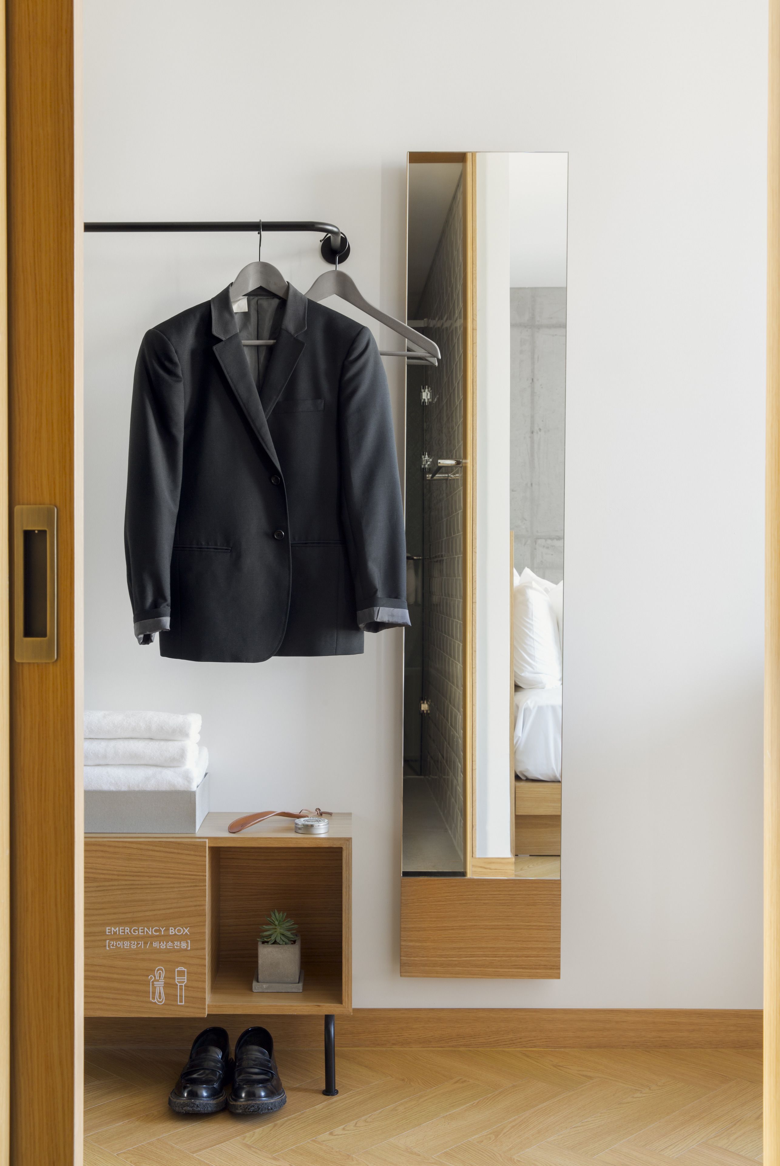A neatly organized bedroom with a hanging coat rack, a bedside table with a plant, and a visible emergency box.