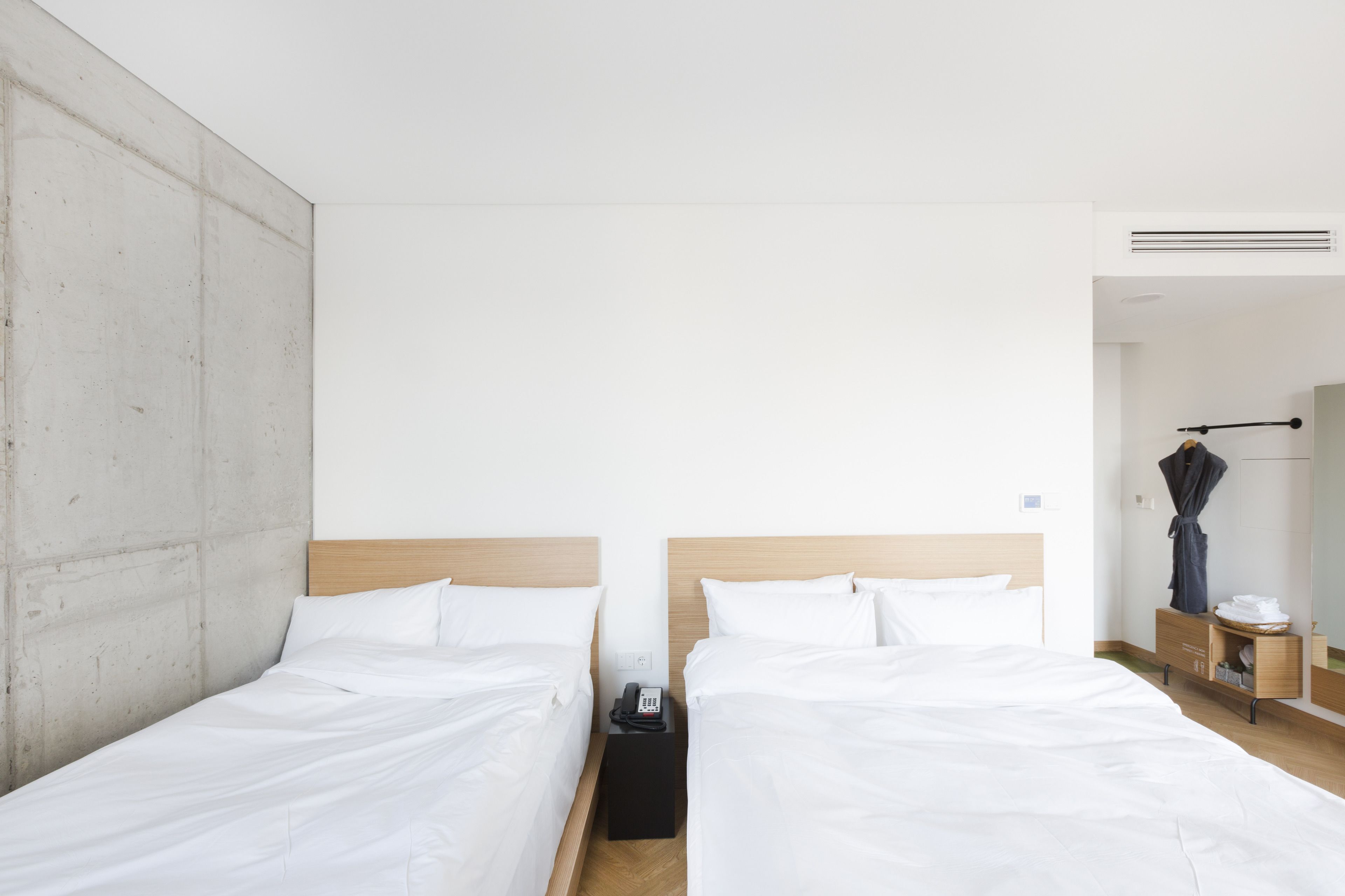 A hotel room with two single beds, a wooden headboard, a small bedside table with a phone, and a robe hanging on a rack.