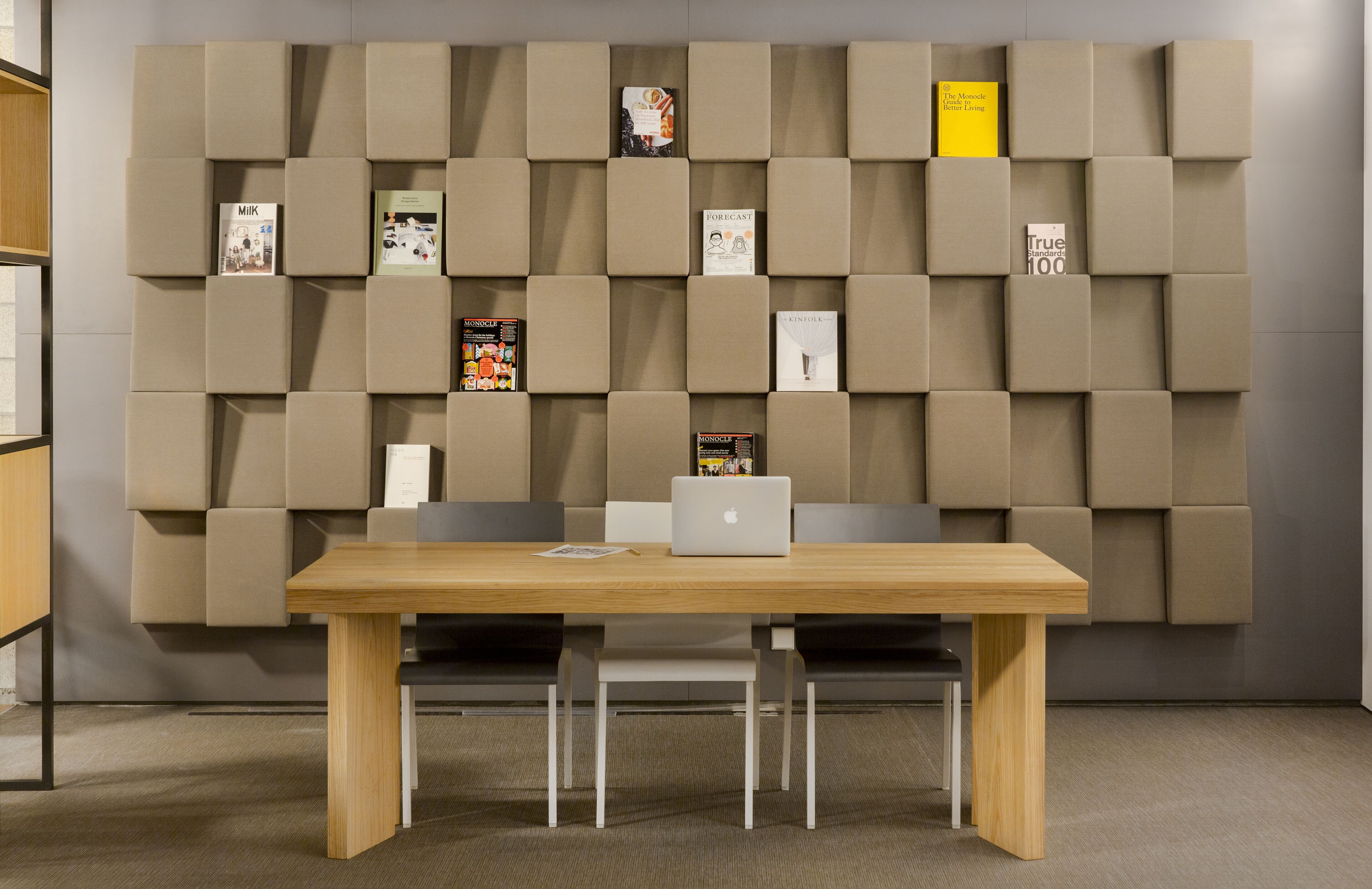 A modern office space with a wooden table, two white chairs, and a wall-mounted bookshelf with various books and a laptop.