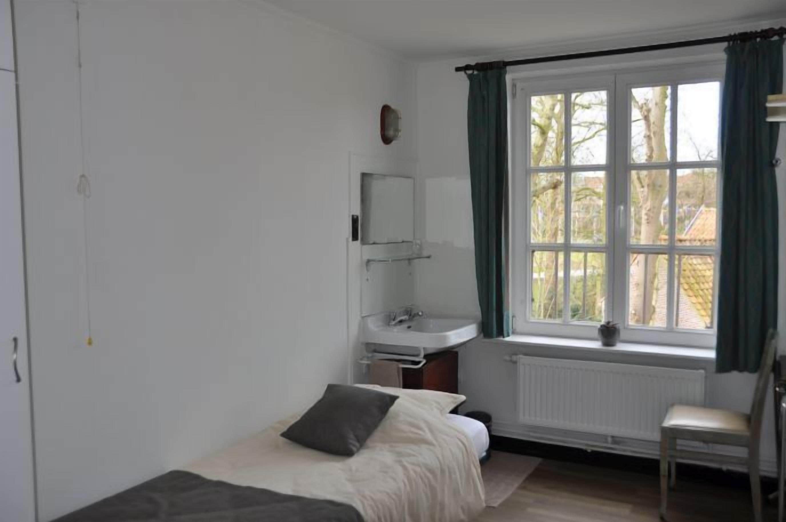 A bedroom with a bed, a bedside table, a chair, a window with curtains, a radiator, and a small shelf with a plant.