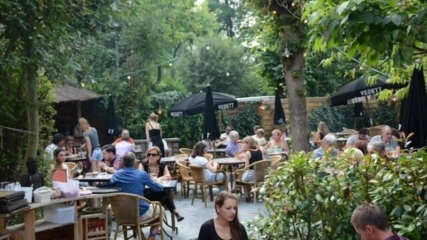 Un café en plein air, avec des tables et des chaises, entouré de verdure.