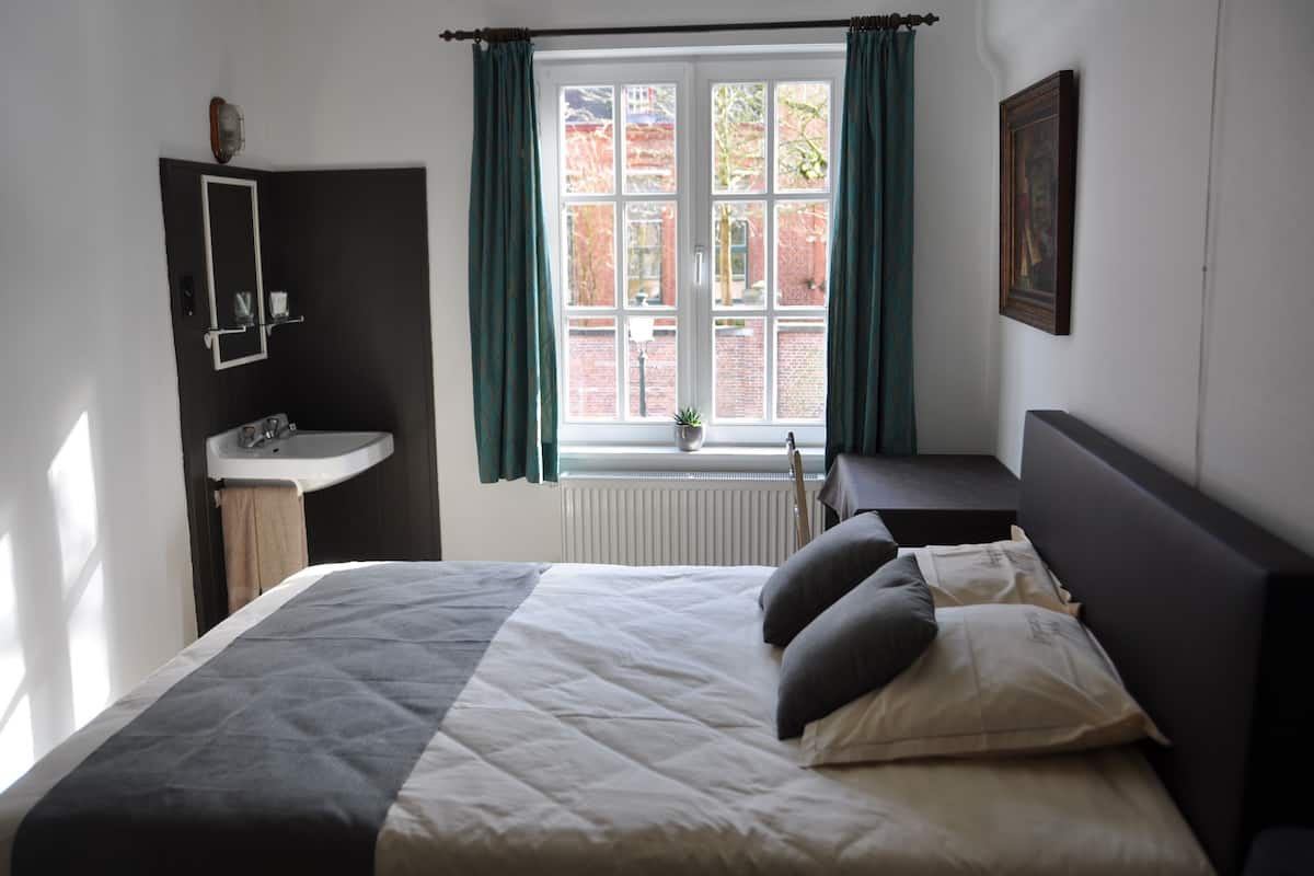 Une chambre à coucher comprenant un lit, une table de chevet, une fenêtre avec des rideaux, un radiateur et un miroir fixé au mur.