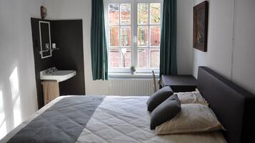 Un dormitorio con una cama, una mesita de noche, una ventana con cortinas, un radiador y un espejo fijado a la pared.