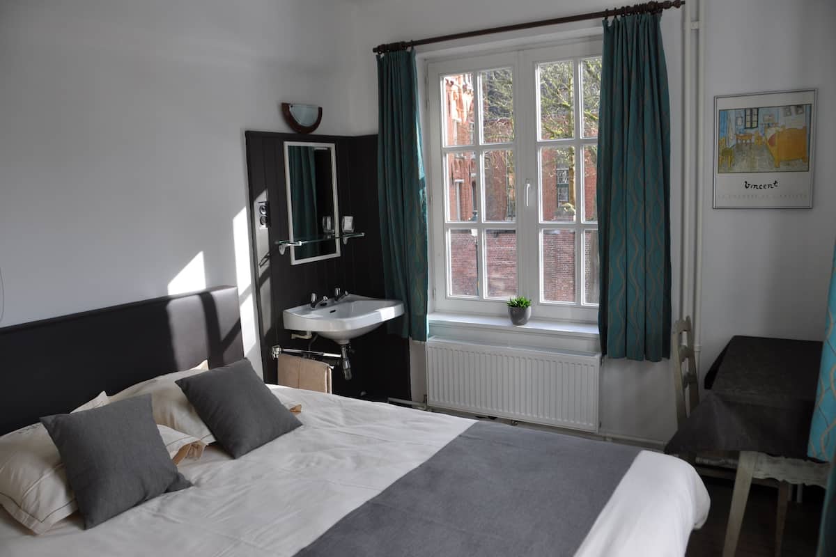 Une chambre à coucher avec un lit, une fenêtre avec des rideaux, une table de chevet, un miroir et un radiateur.