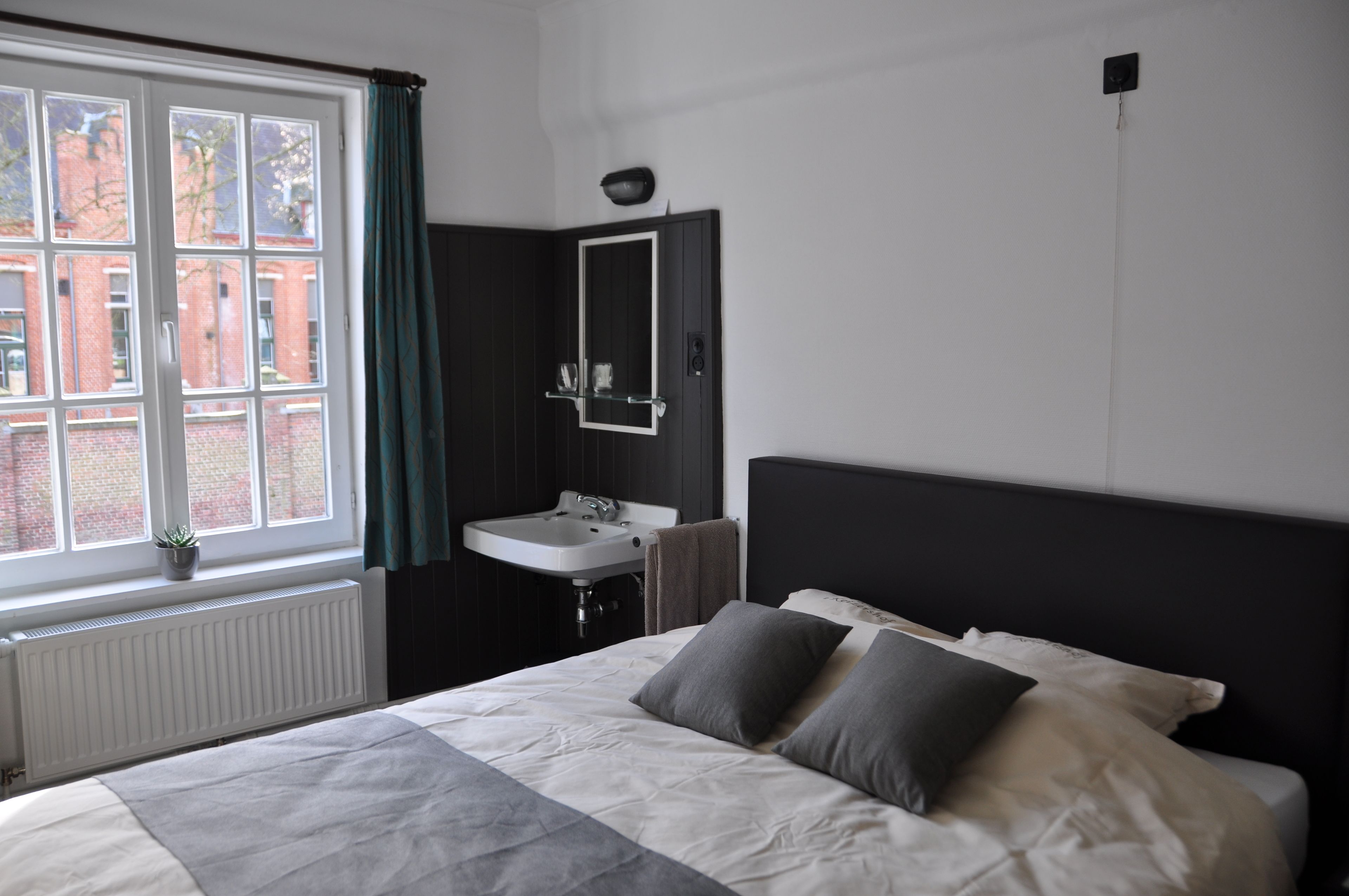 A bedroom with a large bed, a window with curtains, a bedside table, and a wall-mounted mirror.