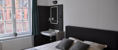 A bedroom with a large bed, a window with curtains, a bedside table, and a wall-mounted mirror.