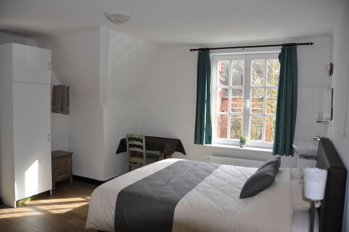 Une chambre à coucher comprenant un lit, une armoire, une petite table avec une chaise, une fenêtre avec des rideaux et un radiateur.