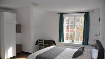 Un dormitorio con cama, armario, una pequeña mesa con silla, ventana con cortinas y radiador.