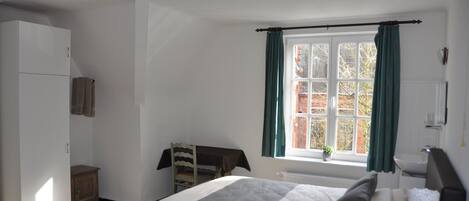 Un dormitorio con cama, armario, una pequeña mesa con silla, ventana con cortinas y radiador.