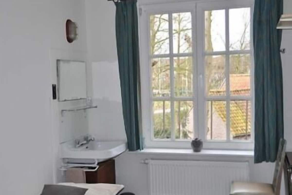 Une chambre à coucher comprenant un lit, une chaise, une fenêtre avec des rideaux, un radiateur et une petite plante posée sur le rebord de la fenêtre.