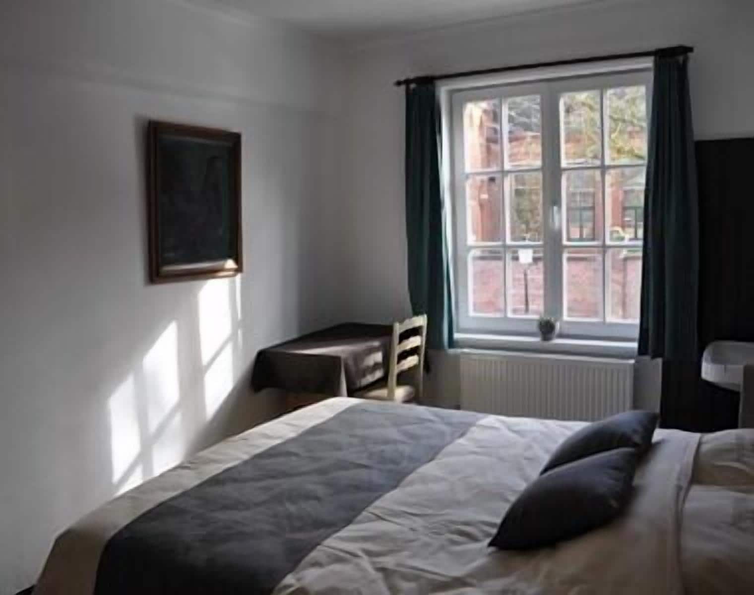Ein Schlafzimmer mit Bett, Sessel, Nachttisch, Fenster mit Vorhängen und einem gerahmten Bild an der Wand.