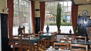 Un espace repas avec des tables et des chaises en bois, un sol à carreaux et de grandes fenêtres avec des rideaux.