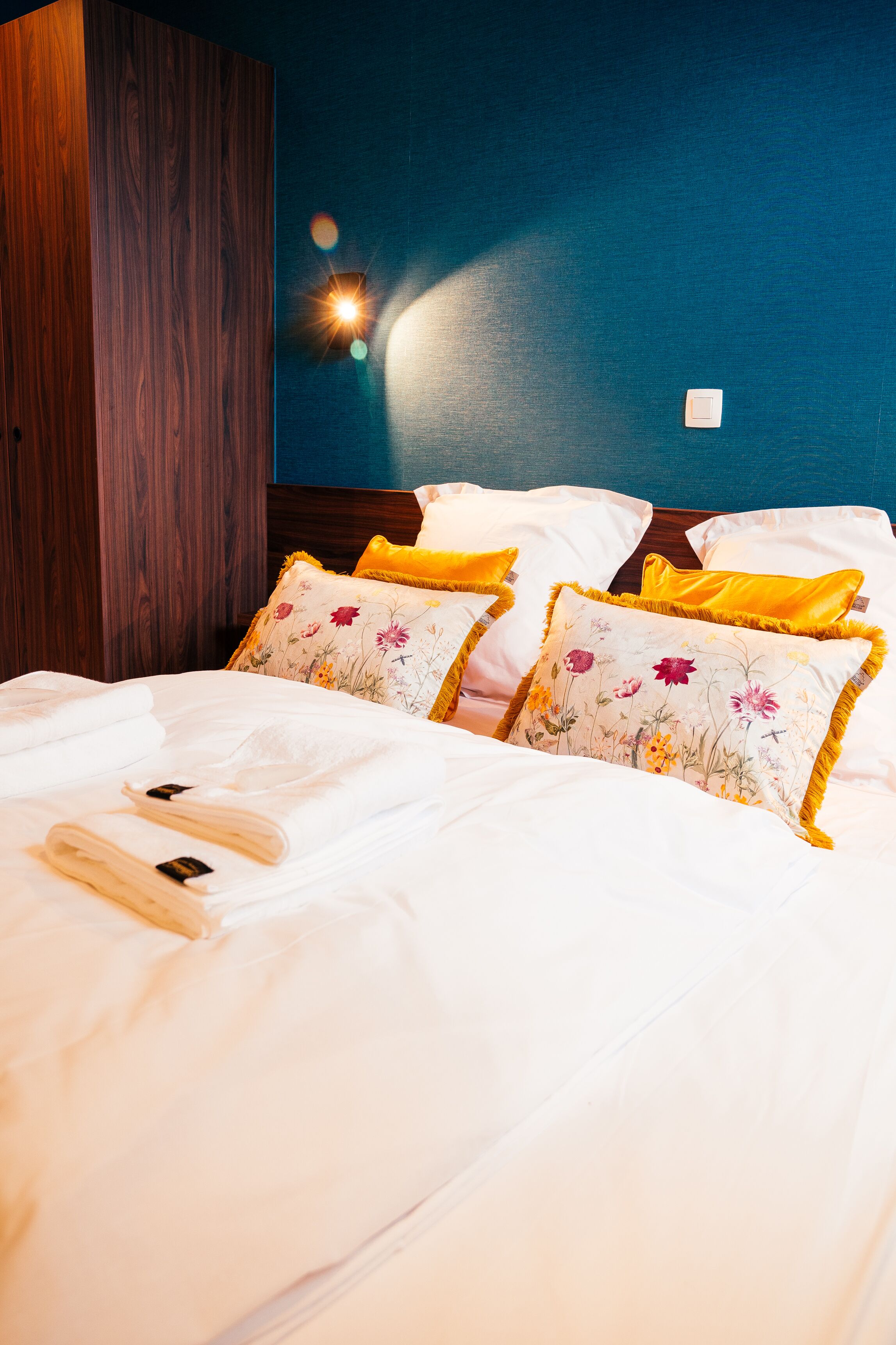 Una cama bien hecha con sábanas blancas y almohadones florales, un armario de madera y una lámpara fijada a la pared.