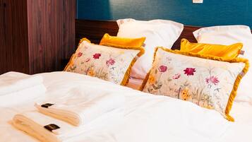 Una cama bien hecha con sábanas blancas y almohadones florales, un armario de madera y una lámpara fijada a la pared.