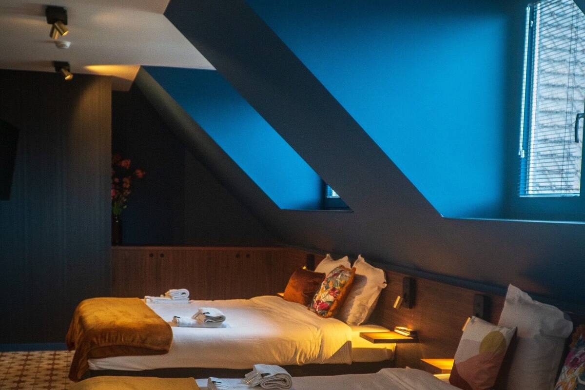 A hotel room with two beds, a sloped ceiling, and a large window with blinds.